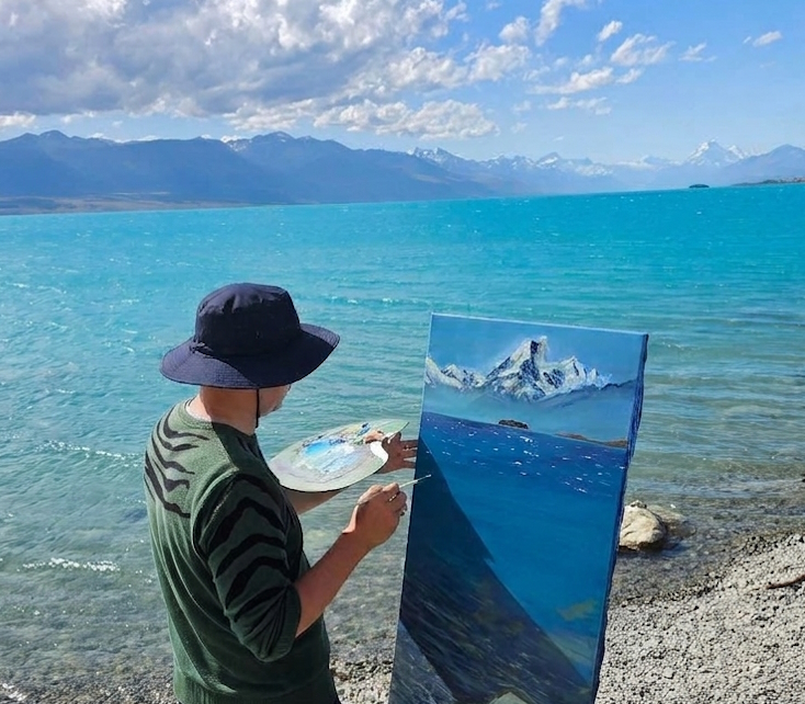 Anton painting a snow-capped mountain in oils, plein air beside a turquoise alpine lake.
